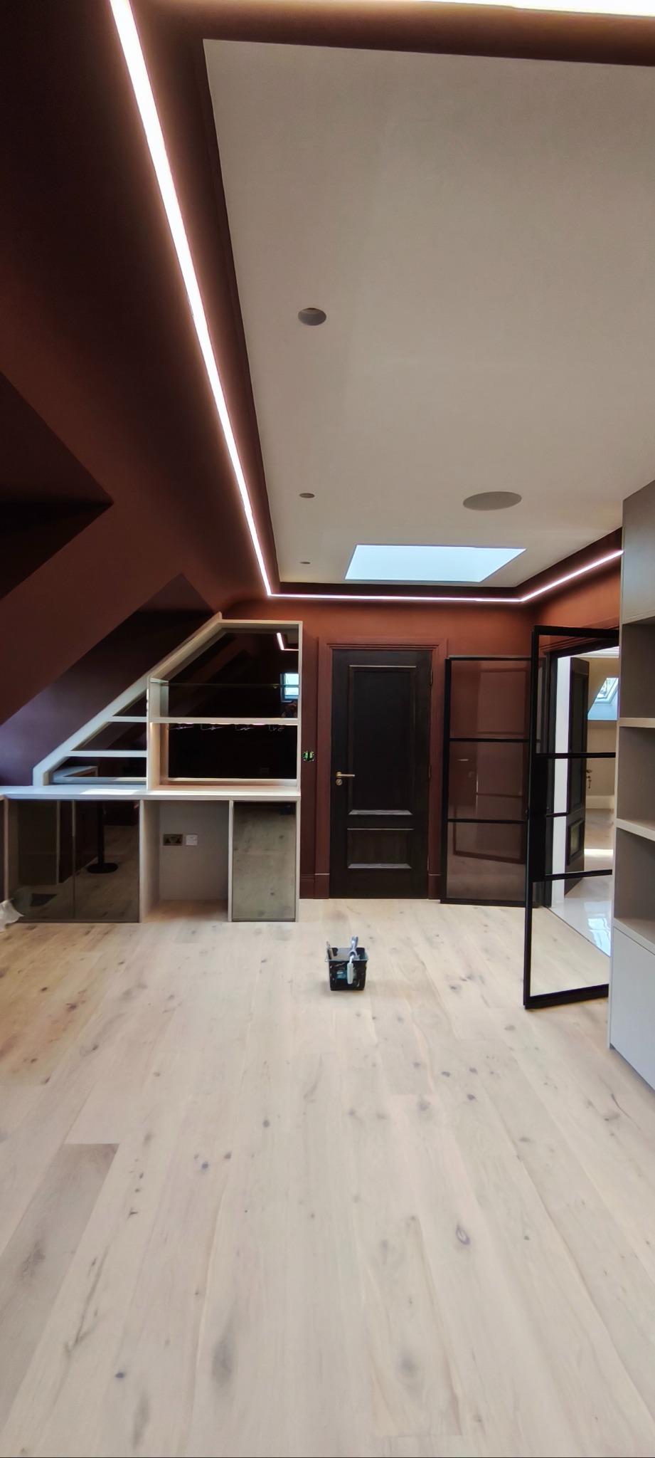 Wykończone wnętrze z drewnianą podłogą, zabudową pod skosem i oświetleniem LED. Widoczny pojemnik z narzędziami malarskimi na podłodze.