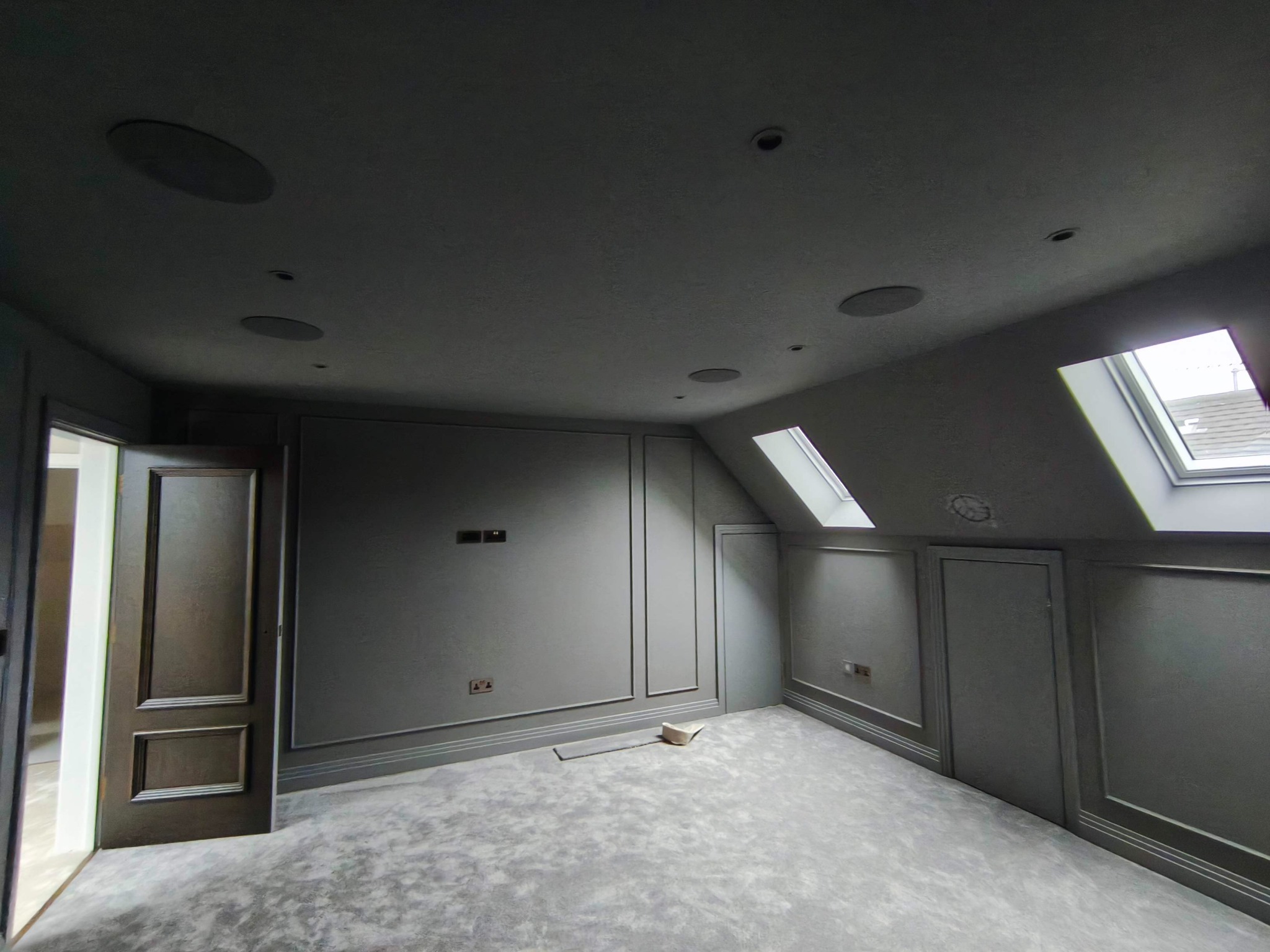 Pokój na poddaszu po remoncie: szare ściany z dekoracyjnymi listwami, okna dachowe, szara wykładzina, wbudowane głośniki sufitowe. Widoczne gniazdka elektryczne.