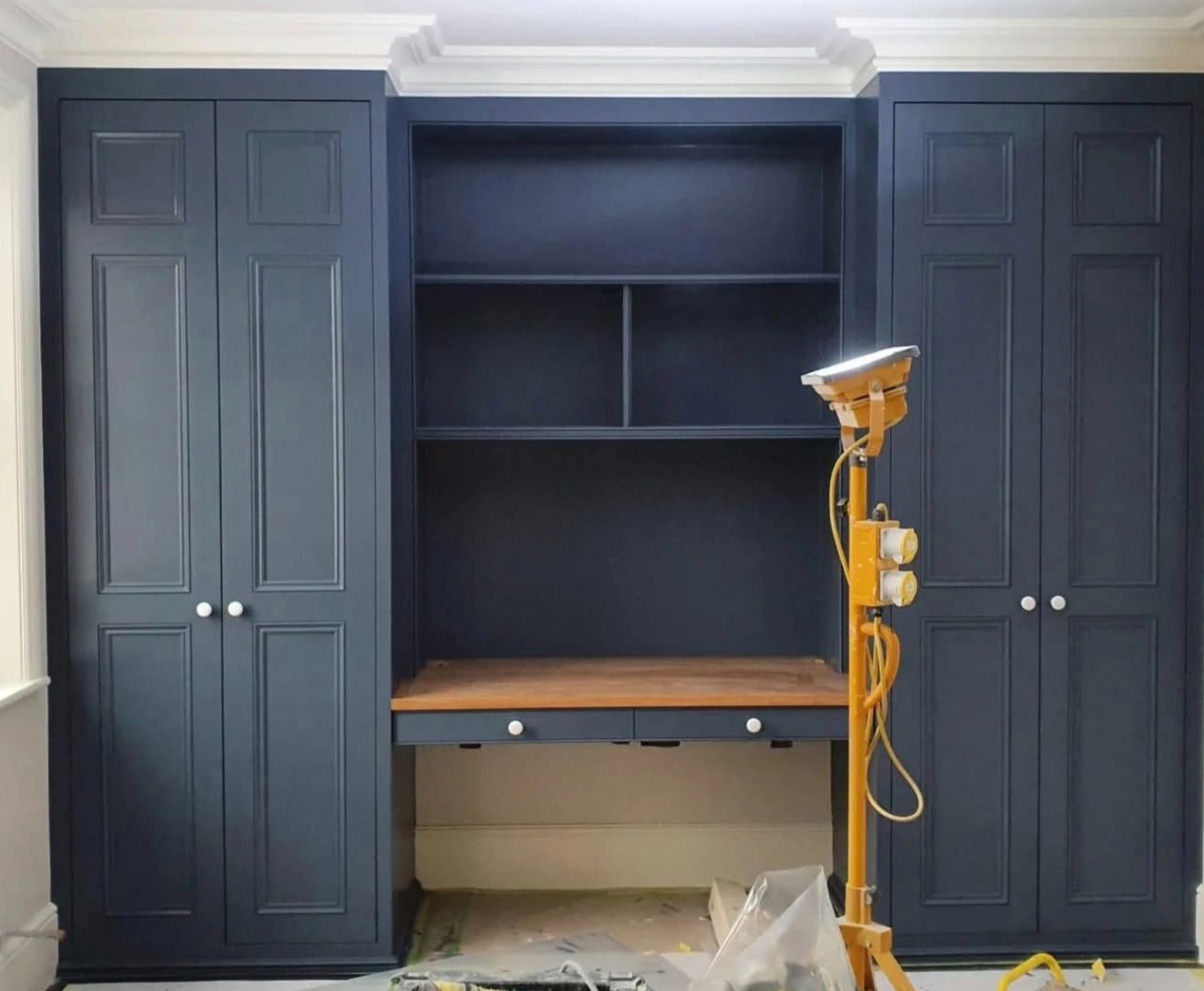 Granatowa zabudowa meblowa z biurkiem i półkami, oświetlona lampą budowlaną. Widoczne ślady prac wykończeniowych na podłodze.