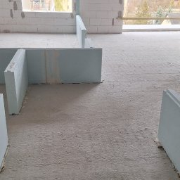 MarcinBud - Wnętrze w trakcie budowy z widocznymi ścianami z bloczków i częściowo zamontowanymi płytami gipsowymi, palety z płytami MultiGips w tle, surowy beton na podłodze.
