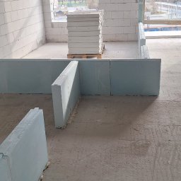 MarcinBud - Wnętrze w trakcie budowy z białymi ścianami z betonu komórkowego i ustawionymi płytami gipsowymi, palety z materiałami budowlanymi w tle.