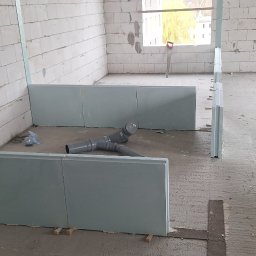 MarcinBud - Wnętrze w trakcie budowy z białymi ścianami z betonu komórkowego, częściowo zamontowaną izolacją podłogową i rurami kanalizacyjnymi. Widoczny poziomica.