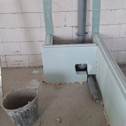 MarcinBud - Niedokończona zabudowa z płyty gipsowej w surowym wnętrzu. Widoczne rury, poziomica i wiadro. Ściany z betonu komórkowego.