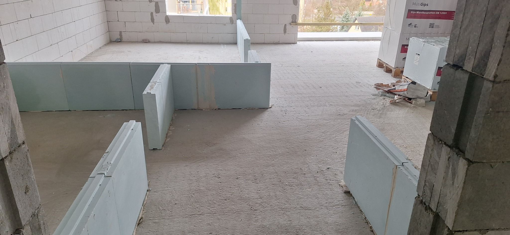 Wnętrze w trakcie budowy z widocznymi ścianami z bloczków i częściowo zamontowanymi płytami gipsowymi, palety z płytami MultiGips w tle, surowy beton na podłodze.