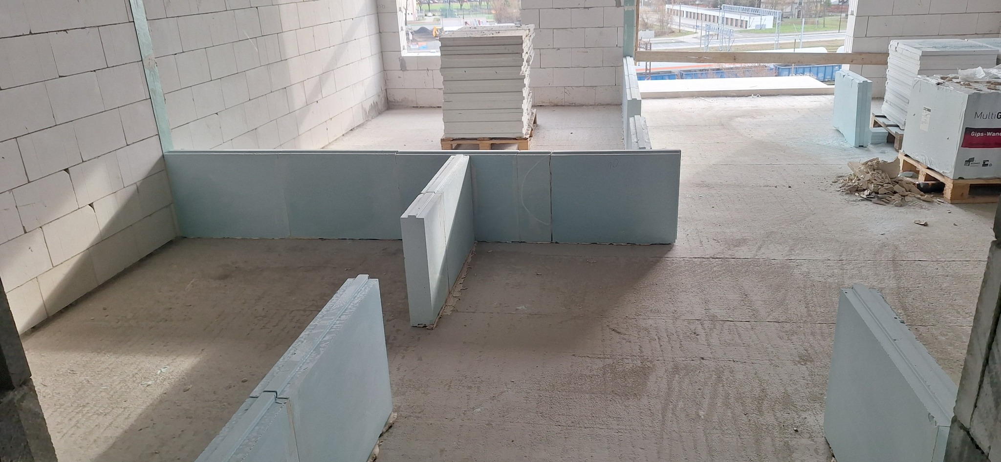 Wnętrze w trakcie budowy z białymi ścianami z betonu komórkowego i ustawionymi płytami gipsowymi, palety z materiałami budowlanymi w tle.