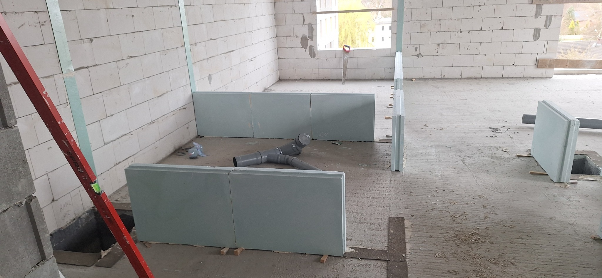 Wnętrze w trakcie budowy z białymi ścianami z betonu komórkowego, częściowo zamontowaną izolacją podłogową i rurami kanalizacyjnymi. Widoczny poziomica.