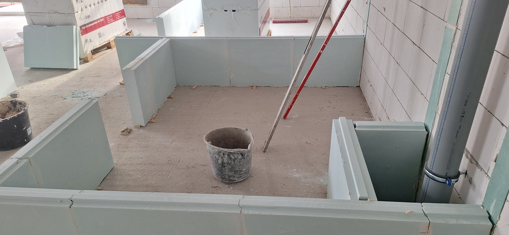 Wnętrze w trakcie budowy z widocznymi ścianami z płyt gipsowych, poziomica oparta o ścianę i wiadro na podłodze. Widoczne materiały budowlane w tle.