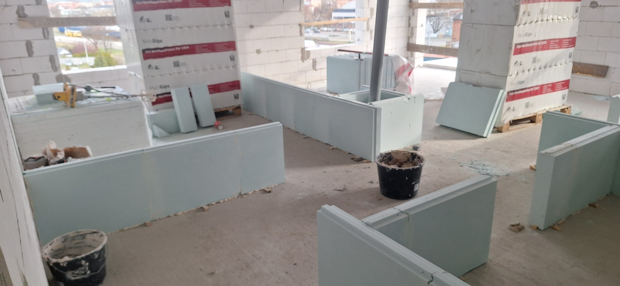 Wnętrze w trakcie montażu ścian z płyt gipsowo-kartonowych. Widoczne płyty, narzędzia i wiadro z zaprawą. Surowy stan wykończenia, okna z widokiem na zewnątrz.