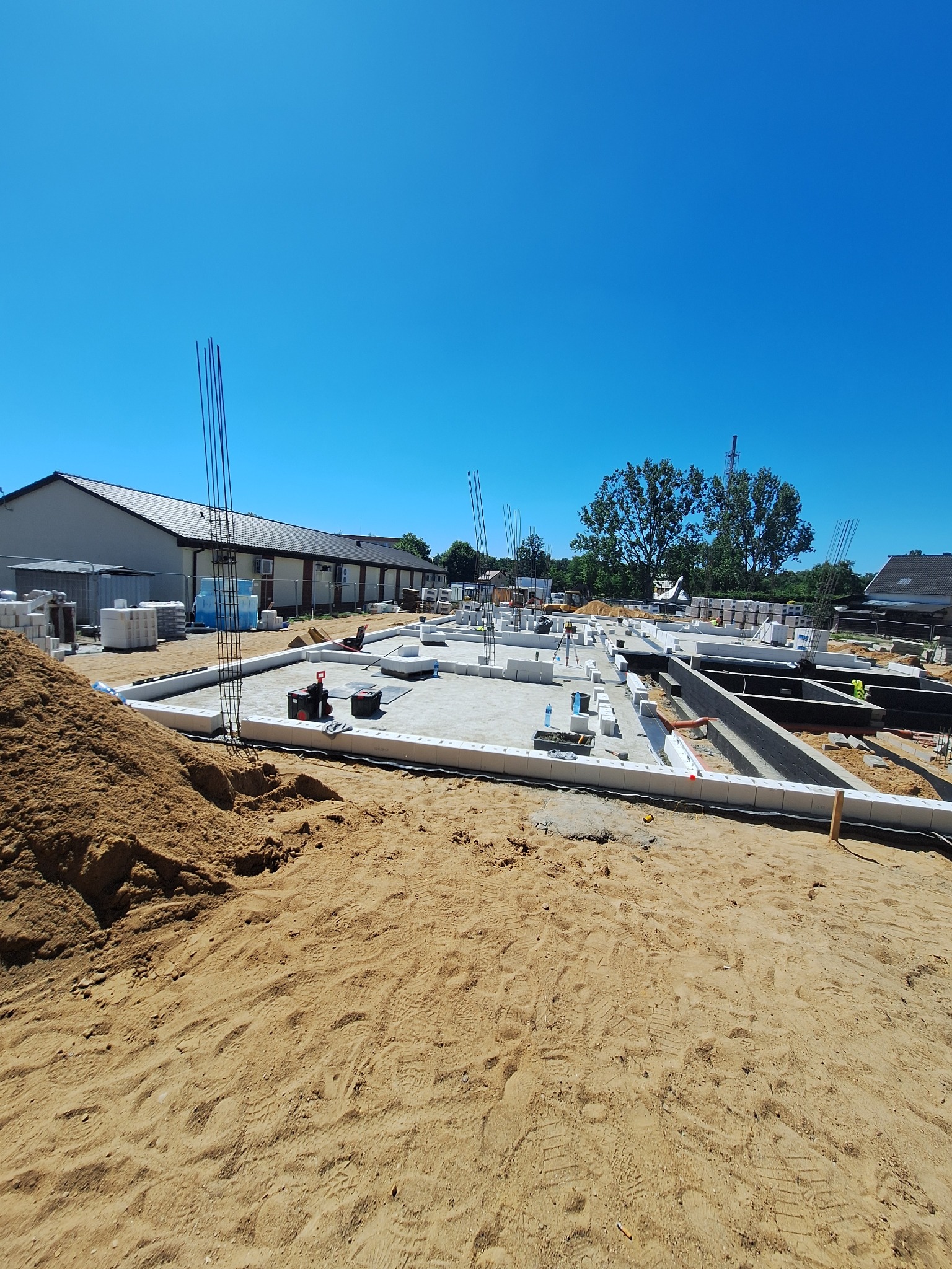 Fundamenty domu w trakcie budowy, widoczne zbrojenia i bloczki betonowe, na pierwszym planie piasek. Słoneczny dzień, błękitne niebo.