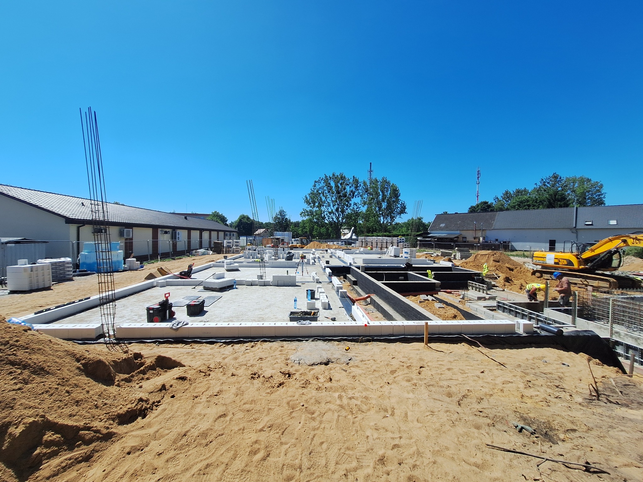 Fundamenty domu w trakcie budowy, widoczne zbrojenia i bloczki fundamentowe. Na budowie obecny jest sprzęt budowlany i pracownicy w odblaskowych kamizelkach.