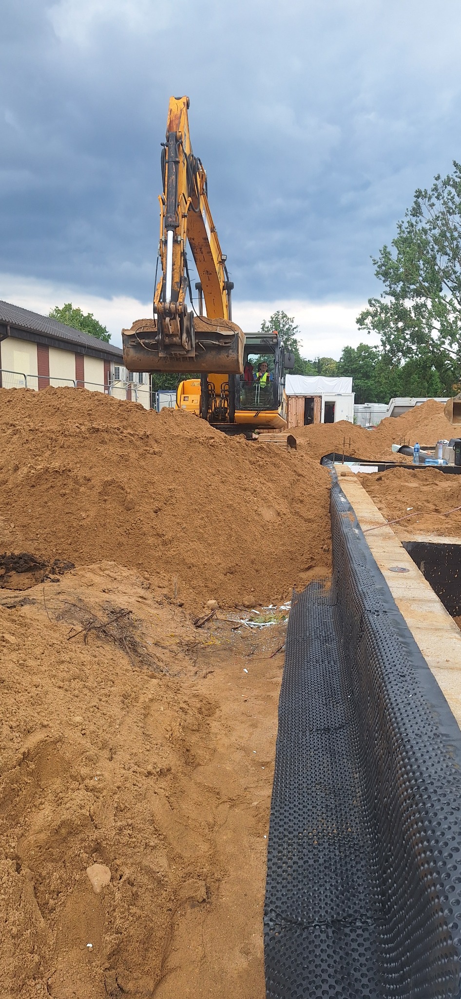 Żółta koparka na placu budowy, przygotowuje teren pod fundamenty. Widoczny wykop, zabezpieczona ściana wykopu folią kubełkową i sterta piasku. Pochmurne niebo w tle.