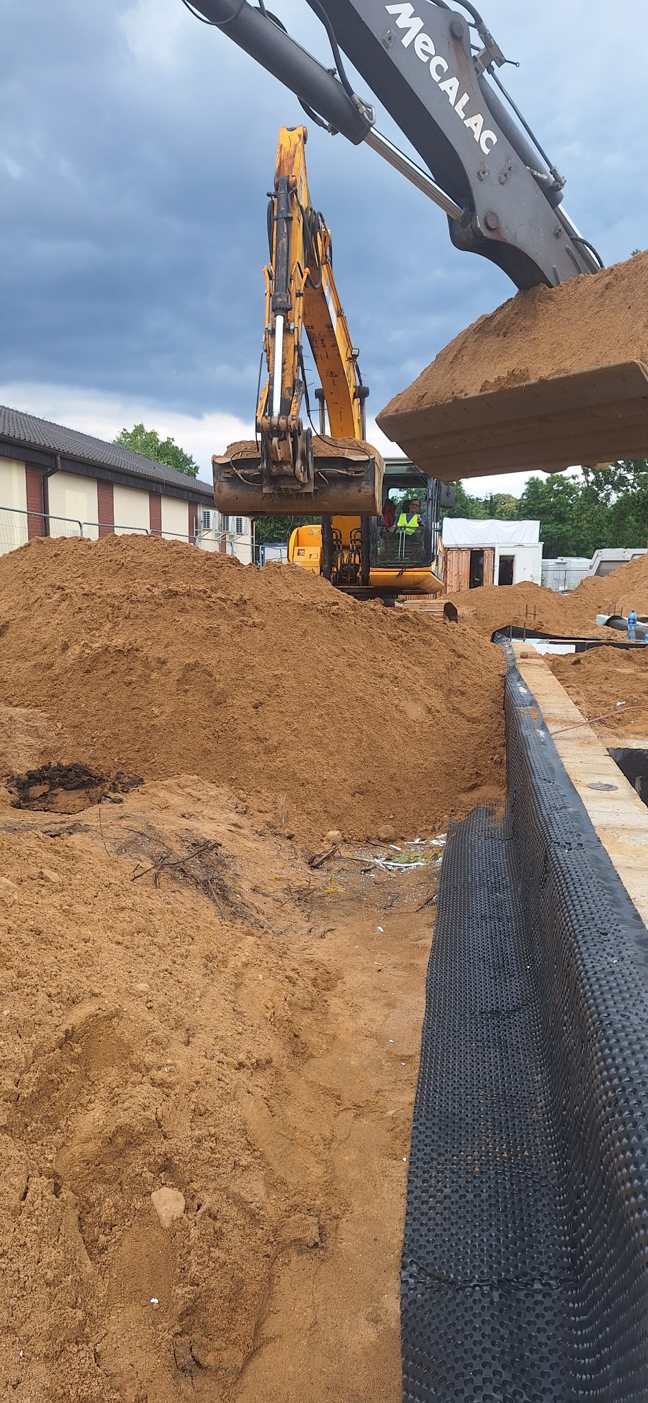 Żółta koparka Mecalac nabiera piasek na budowie fundamentów, widoczna folia kubełkowa i częściowo zarys budynku w tle, pochmurne niebo.