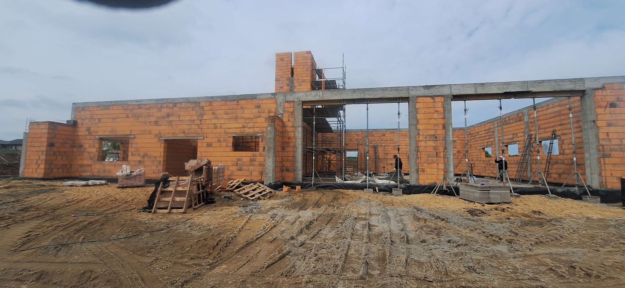 Budowa domu z czerwonej cegły: ściany w trakcie wznoszenia, widoczne otwory okienne i betonowe słupy. Na pierwszym planie piasek i materiały budowlane.