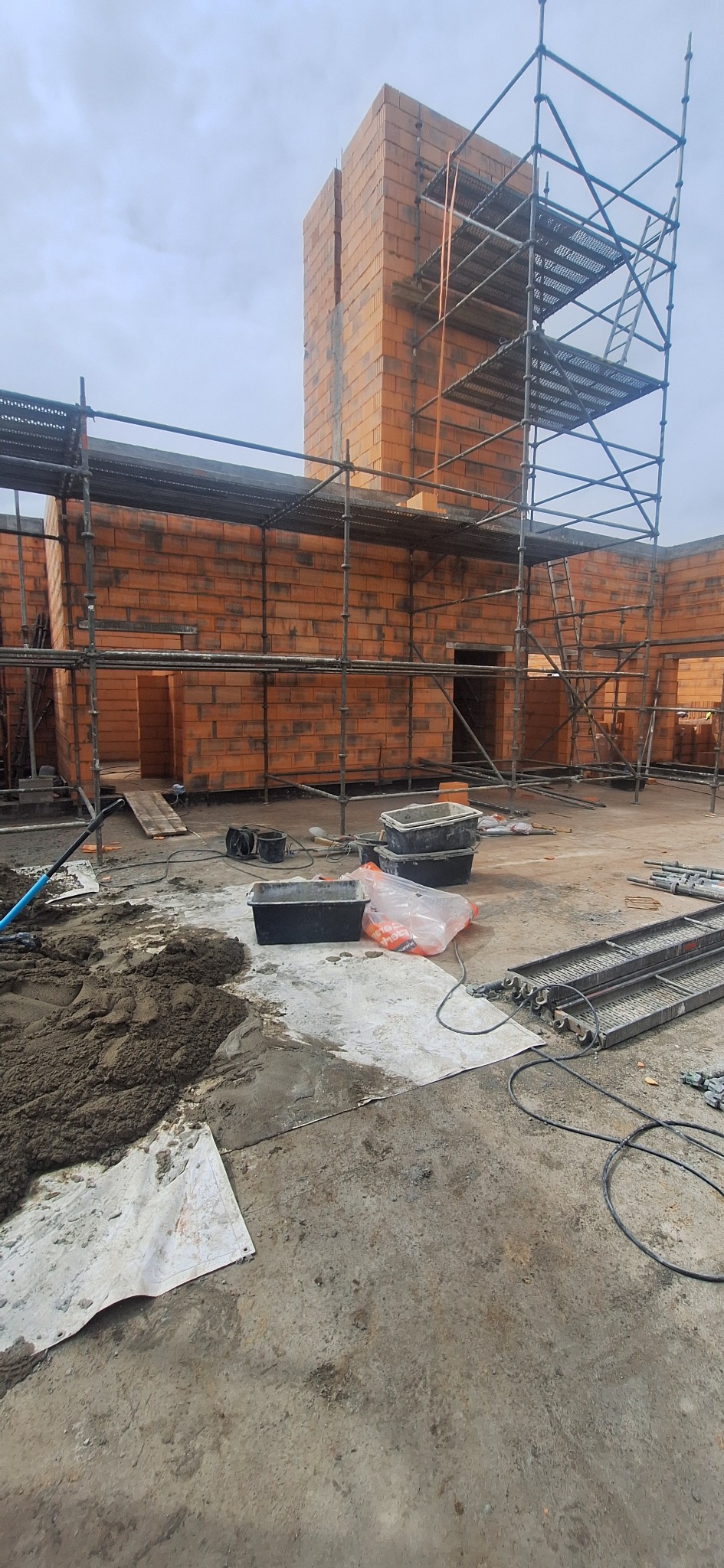 Wznoszenie ścian z czerwonej cegły na budowie domu, widoczne rusztowania i narzędzia murarskie. Ściany w trakcie budowy, z otworami okiennymi i drzwiowymi.