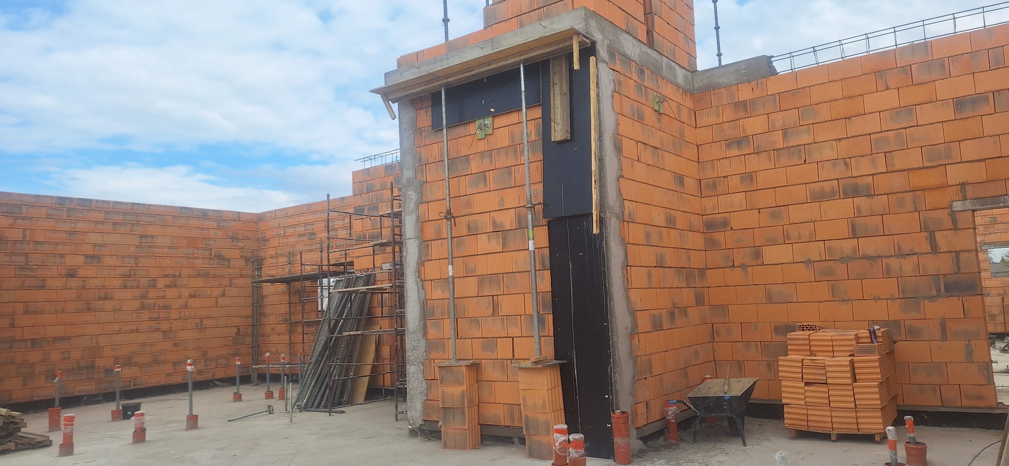 Budowa domu w trakcie: ściany z czerwonej cegły, rusztowania, szalunki pod wylewkę, widoczne materiały budowlane. Etap murowania ścian.