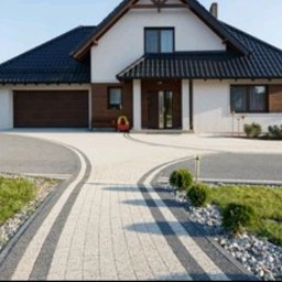Usługi budowlane Piotr Przydacz - Dwukolorowa kostka brukowa tworzy elegancki podjazd do domu z garażem. Widoczne krzewy i trawnik. Jasna elewacja budynku z ciemnym dachem.