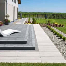 Usługi budowlane Piotr Przydacz - Nowoczesny taras z szarej kostki brukowej, białe płyty chodnikowe, dekoracyjny żwir i roślinność. W tle zielony trawnik i ogrodzenie.