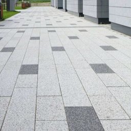 Usługi budowlane Piotr Przydacz - Ułożona, jasnoszara kostka brukowa z ciemnoszarymi wstawkami, tworząca wzór na chodniku przed budynkiem z szarymi panelami. Widok z perspektywy.