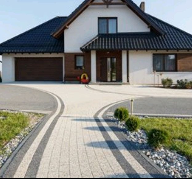 Dwukolorowa kostka brukowa tworzy elegancki podjazd do domu z garażem. Widoczne krzewy i trawnik. Jasna elewacja budynku z ciemnym dachem.