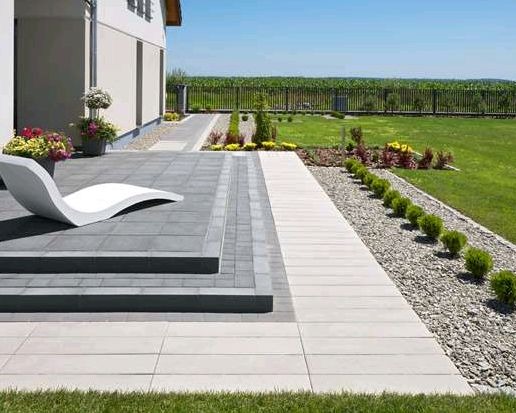 Nowoczesny taras z szarej kostki brukowej, białe płyty chodnikowe, dekoracyjny żwir i roślinność. W tle zielony trawnik i ogrodzenie.
