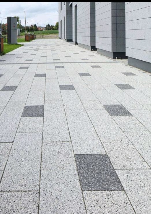 Ułożona, jasnoszara kostka brukowa z ciemnoszarymi wstawkami, tworząca wzór na chodniku przed budynkiem z szarymi panelami. Widok z perspektywy.