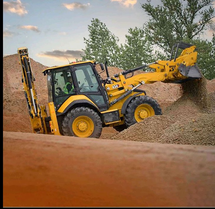 Żółta koparko-ładowarka w trakcie prac ziemnych, wysypująca żwir z łyżki. Widoczny operator w kabinie i fragment drzewa w tle. Ujęcie z dołu.