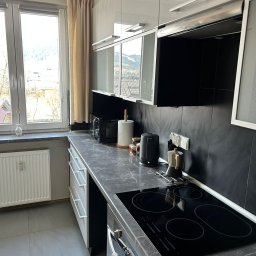 KARPIEL SHINE SERVICES SPÓŁKA Z OGRANICZONĄ ODPOWIEDZIALNOŚCIĄ - Uporządkowana kuchnia z czarnymi płytkami, białymi szafkami i widokiem na góry za oknem. Na blacie czajnik, ekspres do kawy i mikrofalówka. Czyste, szare płytki na podłodze.