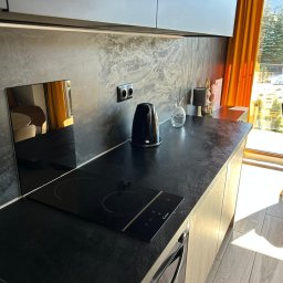 KARPIEL SHINE SERVICES SPÓŁKA Z OGRANICZONĄ ODPOWIEDZIALNOŚCIĄ - Nowoczesna kuchnia z czarnym blatem i płytą indukcyjną. Na blacie czajnik, dekoracje. W tle okno z pomarańczowymi zasłonami i widok na zewnątrz. Minimalistyczny design.