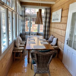 KARPIEL SHINE SERVICES SPÓŁKA Z OGRANICZONĄ ODPOWIEDZIALNOŚCIĄ - Jasne, drewniane wnętrze jadalni z dużym oknem, wiklinowym krzesłem i drewnianym stołem. Widoczne poduszki na ławce i dekoracje na ścianie.