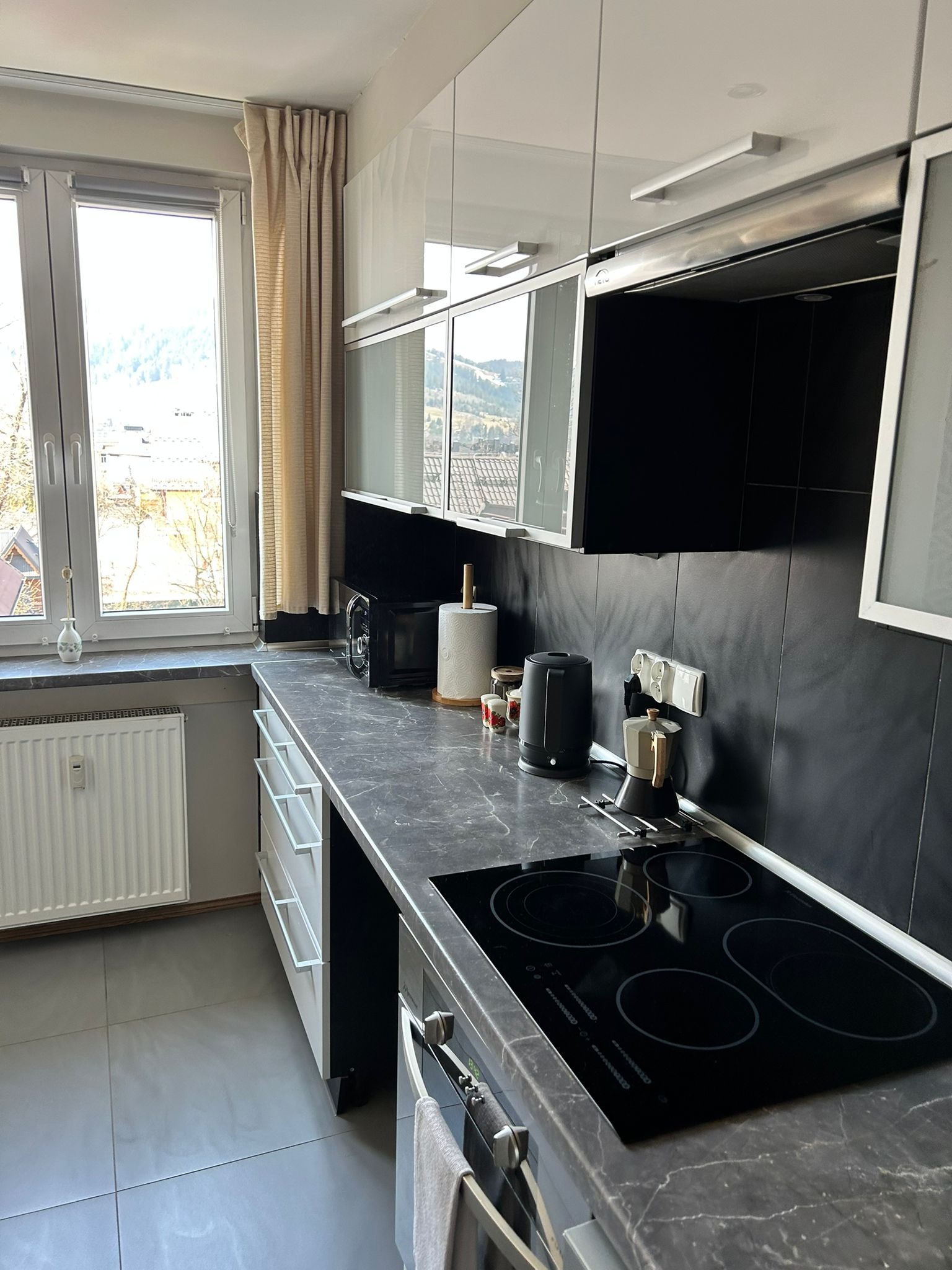 Uporządkowana kuchnia z czarnymi płytkami, białymi szafkami i widokiem na góry za oknem. Na blacie czajnik, ekspres do kawy i mikrofalówka. Czyste, szare płytki na podłodze.