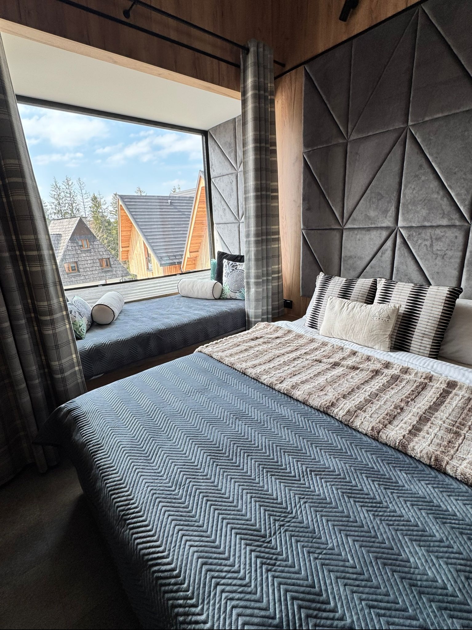 Stylowa sypialnia z dużym oknem i widokiem na góry. Szara tapicerowana ściana z geometrycznym wzorem. Na parapecie poduszki dekoracyjne.