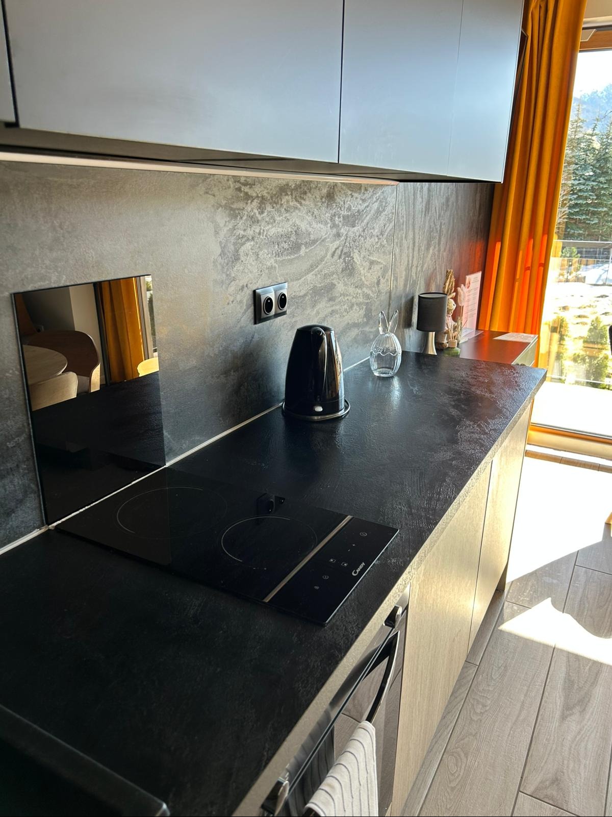 Nowoczesna kuchnia z czarnym blatem i płytą indukcyjną. Na blacie czajnik, dekoracje. W tle okno z pomarańczowymi zasłonami i widok na zewnątrz. Minimalistyczny design.