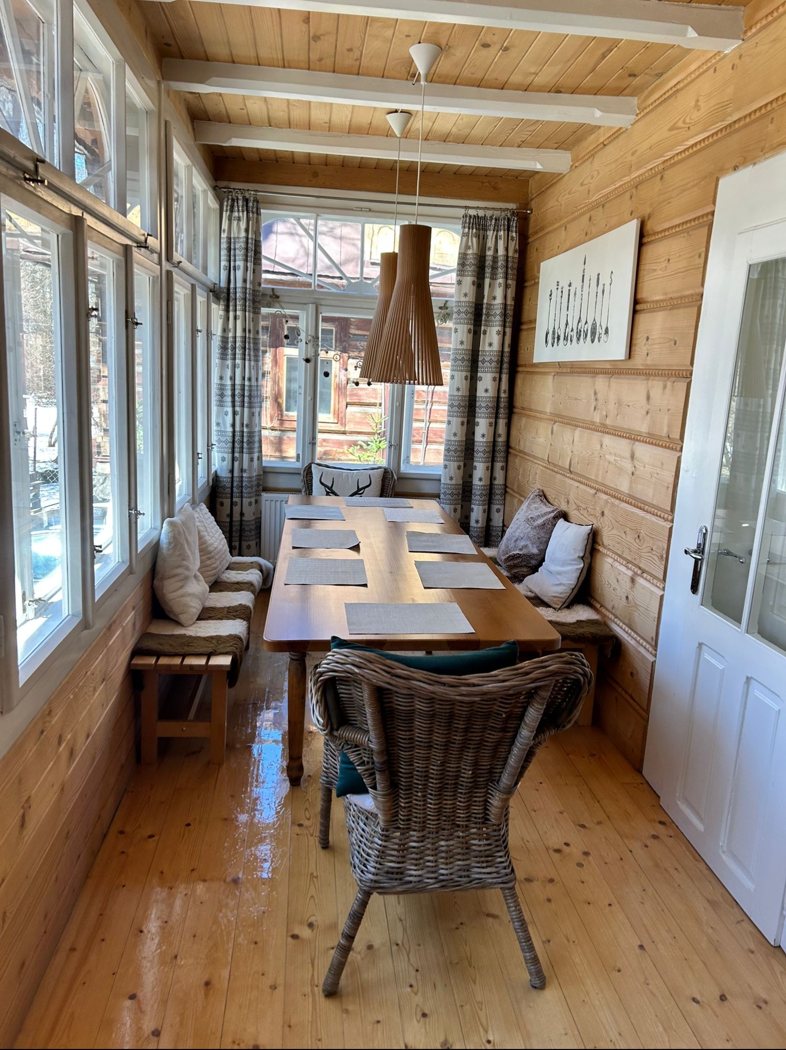 Jasne, drewniane wnętrze jadalni z dużym oknem, wiklinowym krzesłem i drewnianym stołem. Widoczne poduszki na ławce i dekoracje na ścianie.