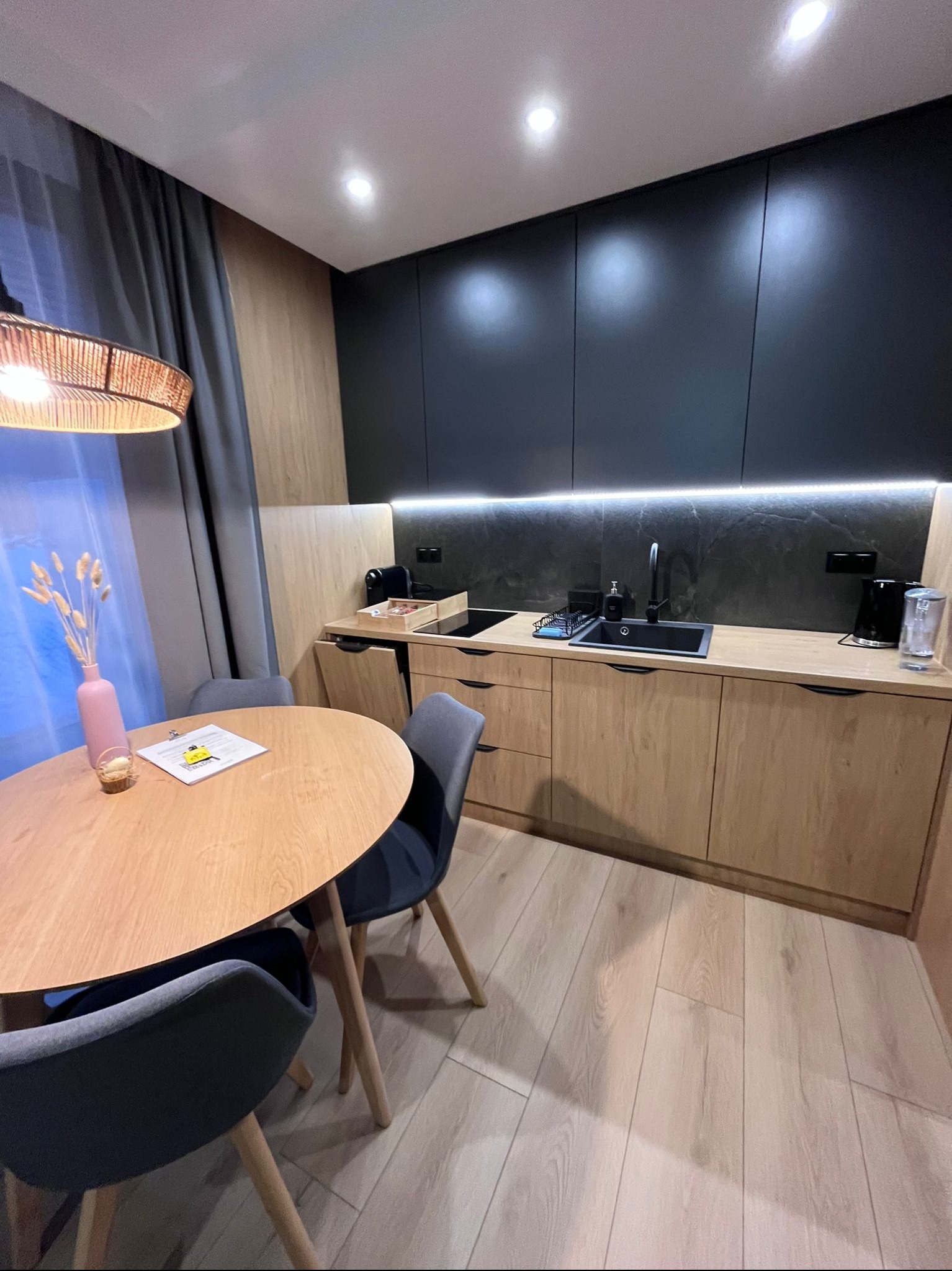 Nowoczesna kuchnia z drewnianymi szafkami i czarnymi frontami, okrągły stół z dekoracją. Minimalistyczny design, jasne drewno na podłodze, subtelne oświetlenie.