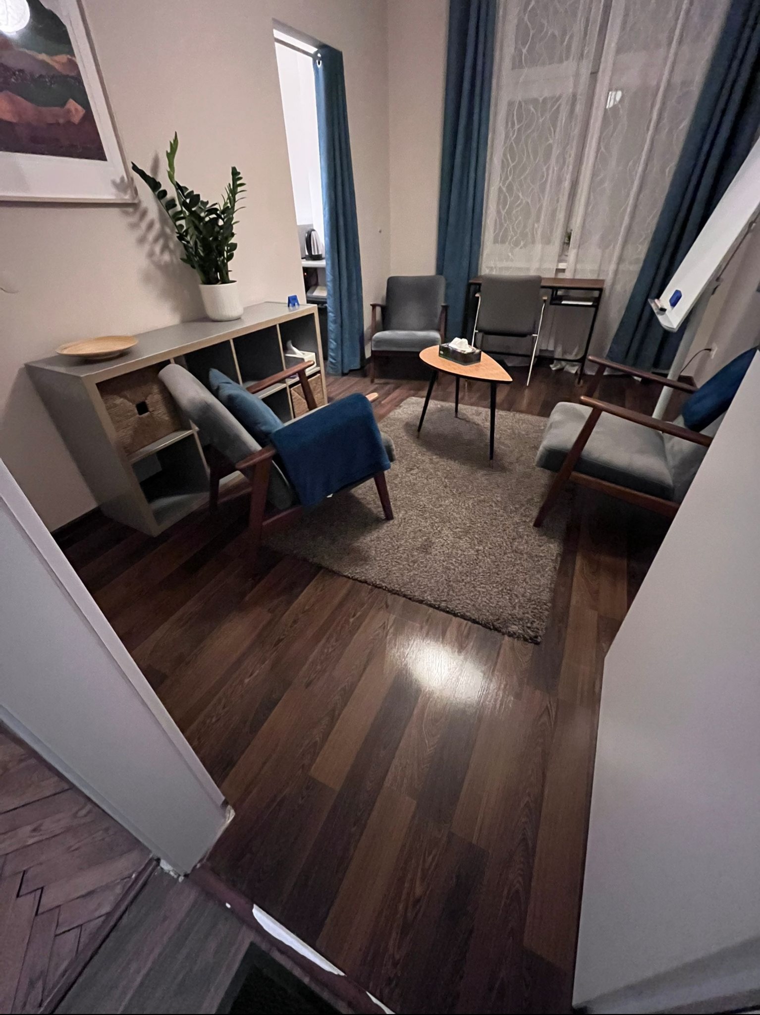 Salon z drewnianą podłogą, szarymi fotelami i zasłonami. Na komodzie roślina w doniczce. Widok zza rogu, wnętrze po sprzątaniu, gotowe do użytku.