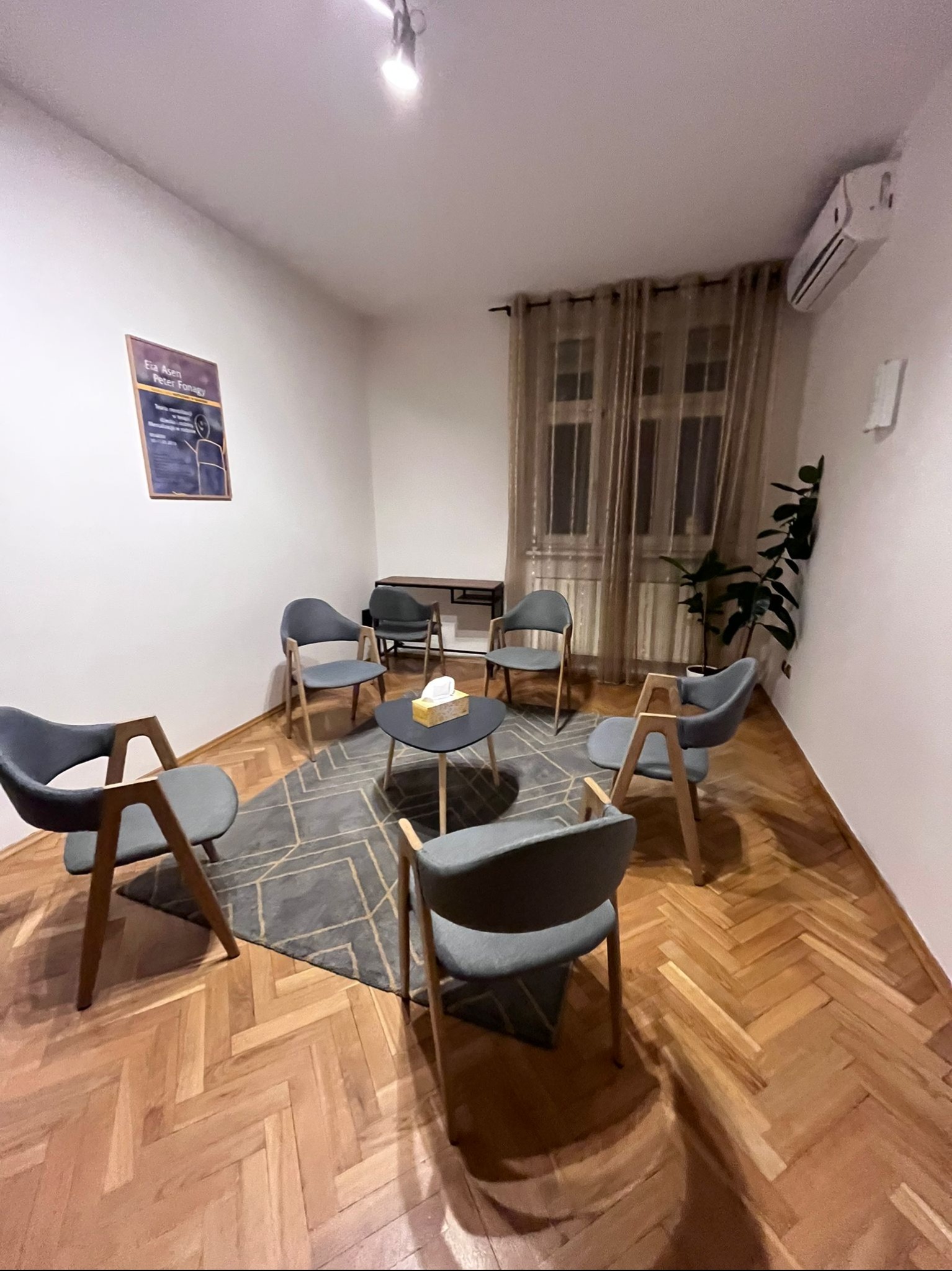 Jasne wnętrze gabinetu z parkietem w jodełkę. Krzesła ustawione w kręgu wokół stolika. Na ścianie plakat i klimatyzator. Roślina doniczkowa przy oknie z zasłonami.