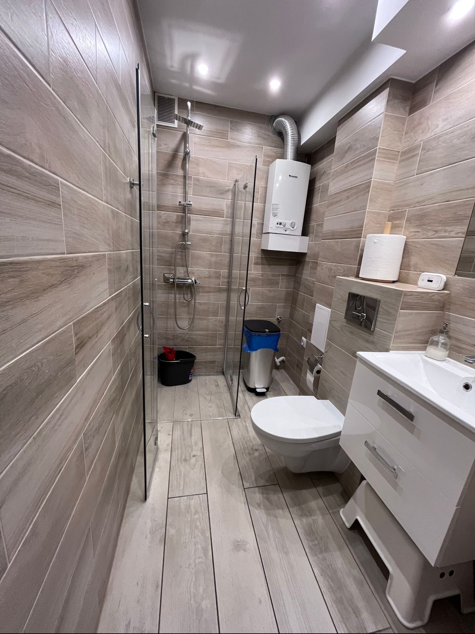 Nowoczesna łazienka po sprzątaniu: kabina prysznicowa, umywalka z szafką, toaleta, bojler. Czyste płytki imitujące drewno na ścianach i podłodze.