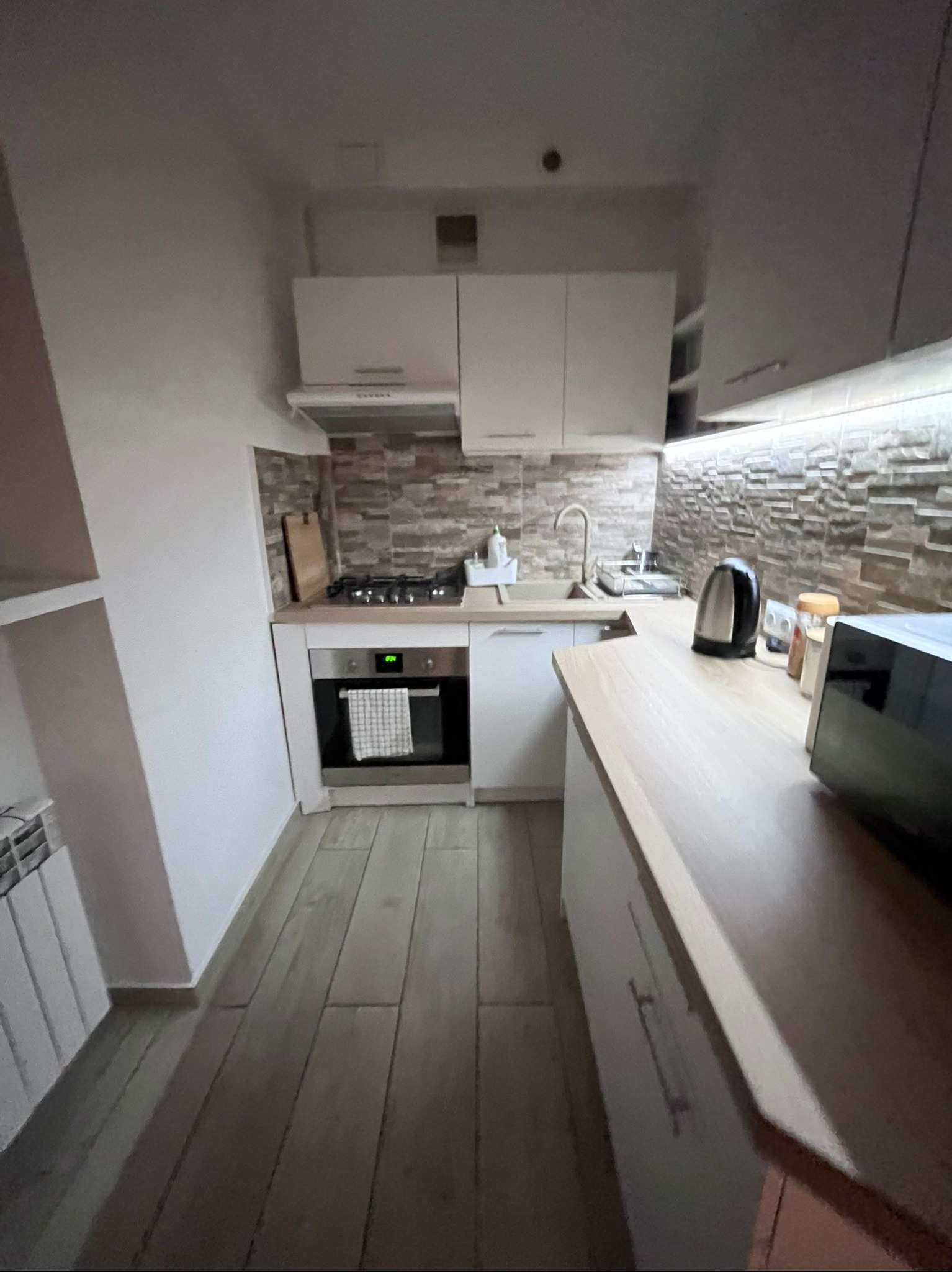 Jasna kuchnia z drewnianym blatem, płytkami imitującymi kamień na ścianie i piekarnikiem z ręcznikiem. Widok na czajnik i mikrofalówkę na blacie.