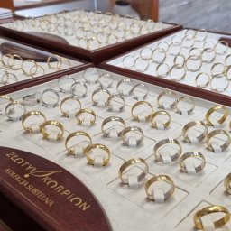 Grawer - Obrączki Legnica - Ekspozycja złotych i srebrnych obrączek ślubnych na eleganckich, drewnianych podkładkach z logo 'Złoty Skorpion'. Różnorodność wzorów i wykończeń.