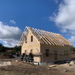 NS-BUD Norbert Szulc - Surowy dom z beżową elewacją w trakcie budowy, z widoczną konstrukcją dachu i rusztowaniami. Błękitne niebo z chmurami w tle.
