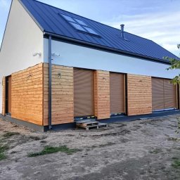 NS-BUD Norbert Szulc - Nowoczesny dom z drewnianą elewacją i roletami zewnętrznymi. Widoczne okna dachowe i fragment ogrodu. Jasna kolorystyka ścian kontrastuje z ciemnym dachem.