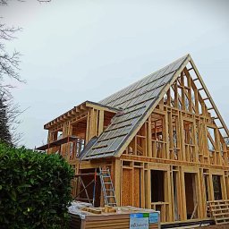 NS-BUD Norbert Szulc - Surowa konstrukcja domu szkieletowego w trakcie budowy, z widocznymi elementami drewnianymi i częściowo ułożonym pokryciem dachu. Na pierwszym planie materiały budowlane.