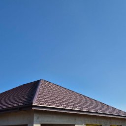 NS-BUD Norbert Szulc - Niski kąt ujęcia na brązowy dach domu w stanie surowym otwartym, na tle błękitnego nieba. Widoczne elementy konstrukcyjne i fragment sąsiedniego budynku.