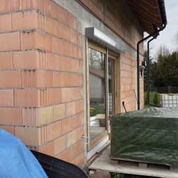 GRYGA Arletta Zawiła - Surowy mur z pustaków ceramicznych z zamontowanym oknem z roletą zewnętrzną w kolorze jasnego drewna. Widok z zewnątrz na budowę domu.