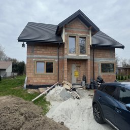 GRYGA Arletta Zawiła - Dom w trakcie budowy z widocznymi oknami w drewnianych ramach, czarnym dachem i stertą piasku przed wejściem. Częściowo widoczny samochód osobowy.
