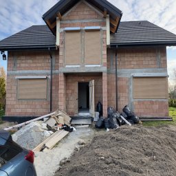 GRYGA Arletta Zawiła - Dom w trakcie budowy z zamontowanymi roletami zewnętrznymi w kolorze beżowym. Widoczne materiały budowlane i worki na śmieci. Częściowo widoczny samochód.