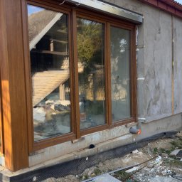 GRYGA Arletta Zawiła - Montaż okien PCV z roletami zewnętrznymi w surowym stanie elewacji. Widoczne brązowe ramy okienne i srebrne kasety rolet. Prace wykończeniowe w toku.