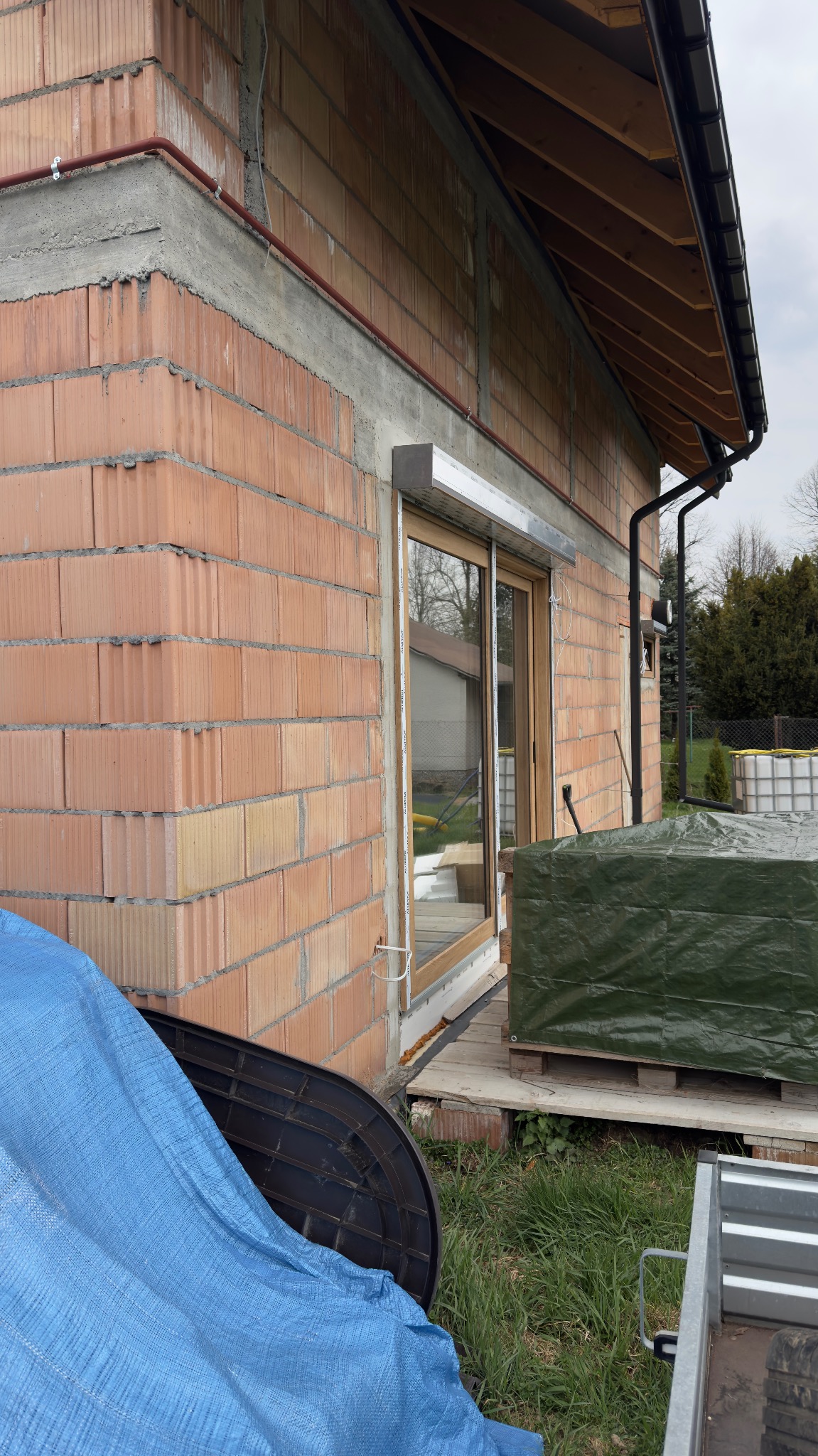 Surowy mur z pustaków ceramicznych z zamontowanym oknem z roletą zewnętrzną w kolorze jasnego drewna. Widok z zewnątrz na budowę domu.