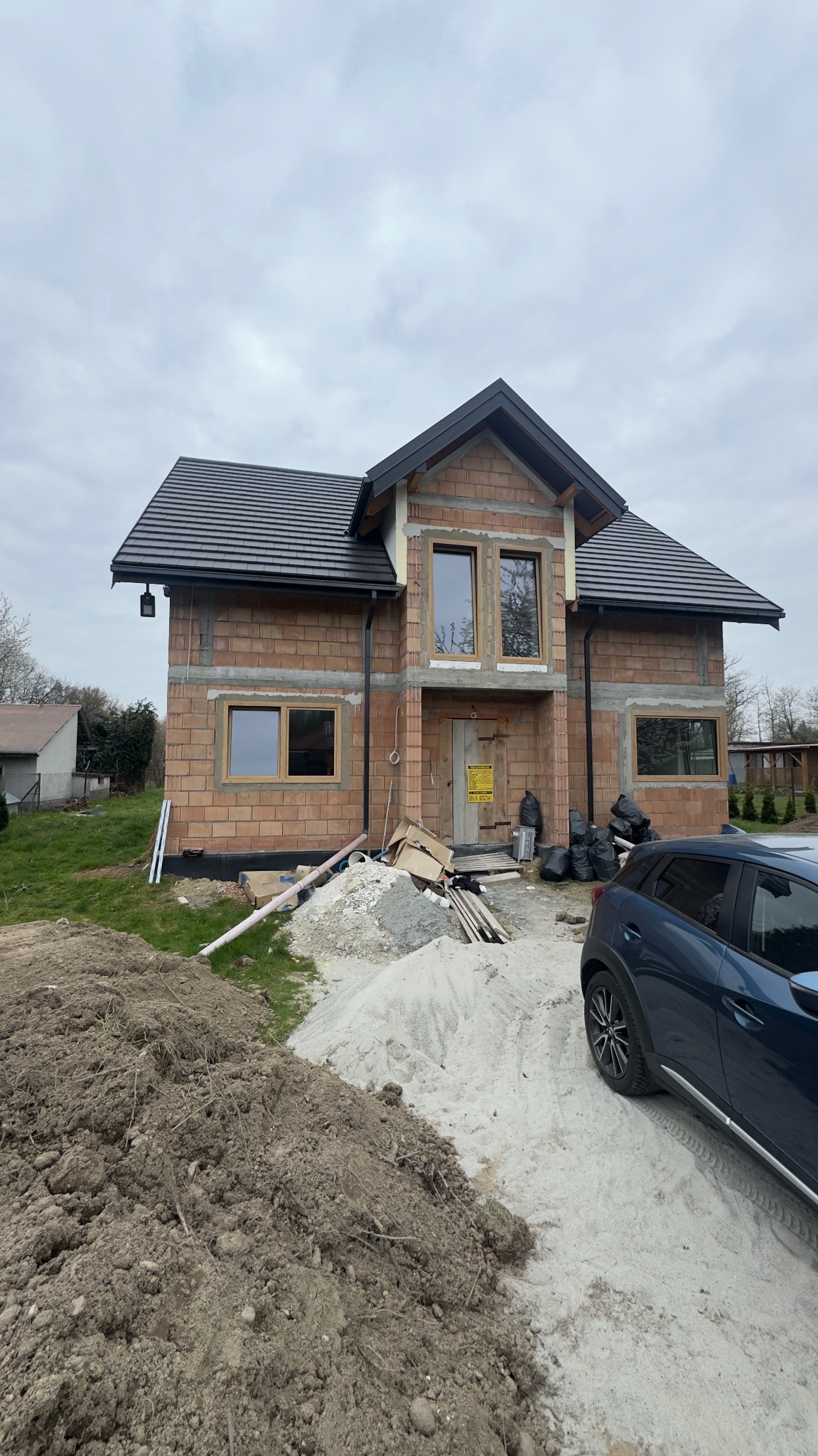 Dom w trakcie budowy z widocznymi oknami w drewnianych ramach, czarnym dachem i stertą piasku przed wejściem. Częściowo widoczny samochód osobowy.