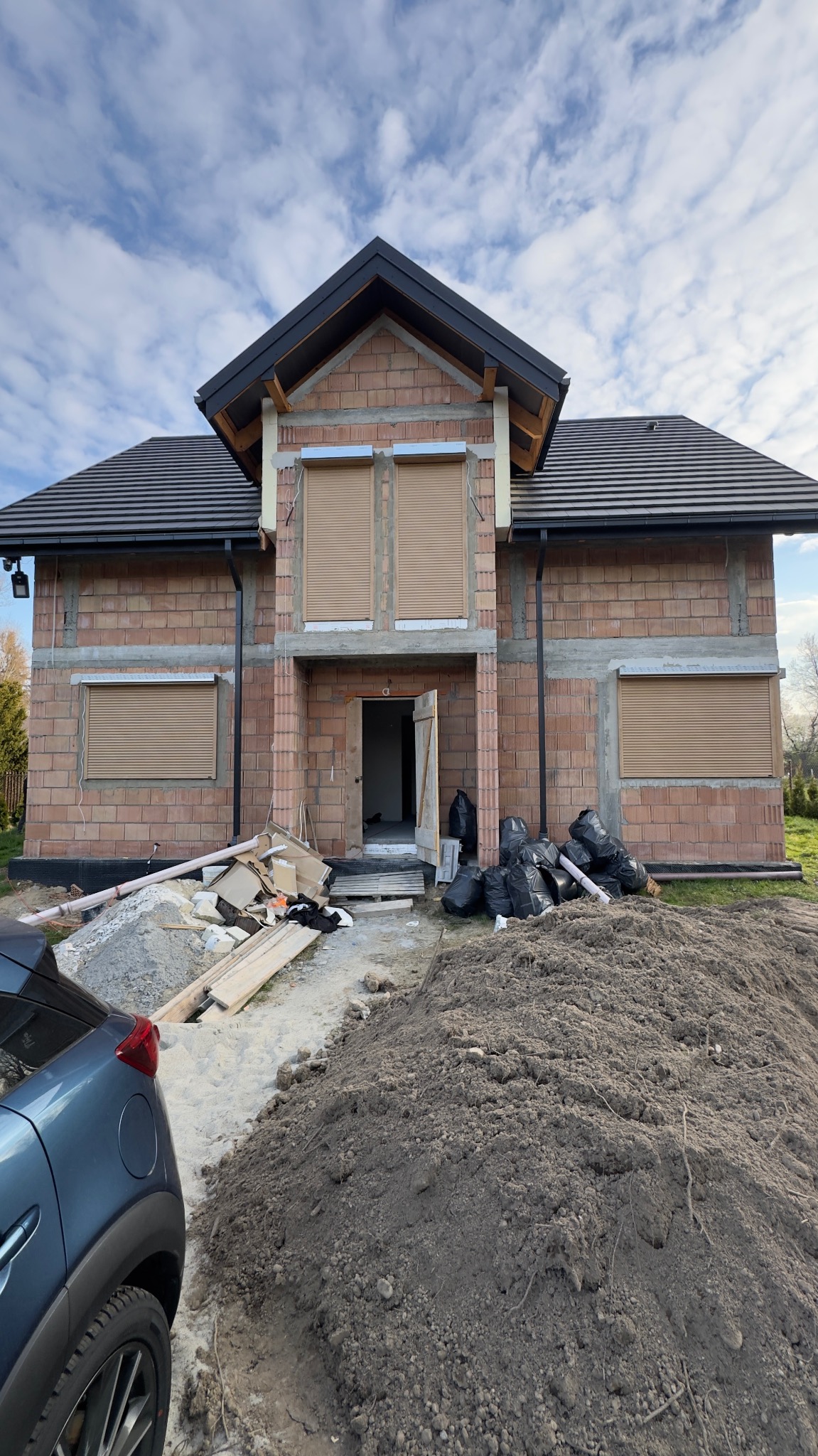 Dom w trakcie budowy z zamontowanymi roletami zewnętrznymi w kolorze beżowym. Widoczne materiały budowlane i worki na śmieci. Częściowo widoczny samochód.